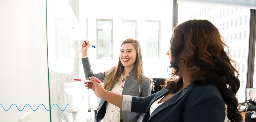 Two businesswomen laugh while collaborating on a dry erase board