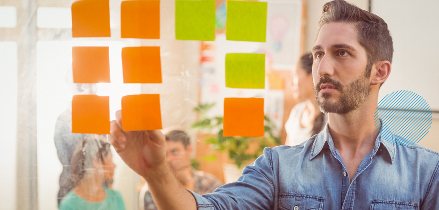 A businessman places sticky notes on a glass wall and colleagues are seated in the background